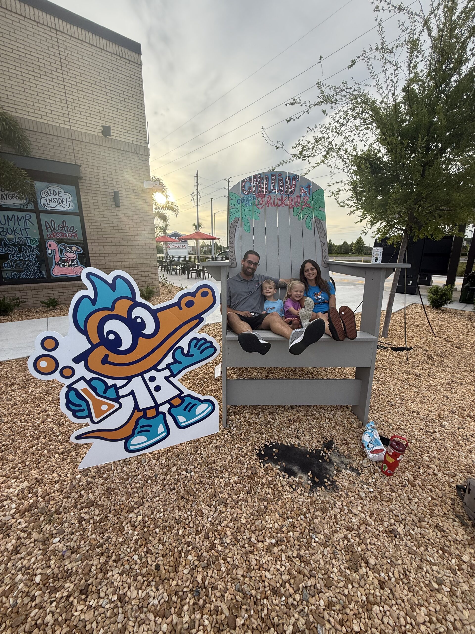 A family seated in an oversized chair outside a Chick-fil-A next to the Formula Plumbing mascot cutout at sunset