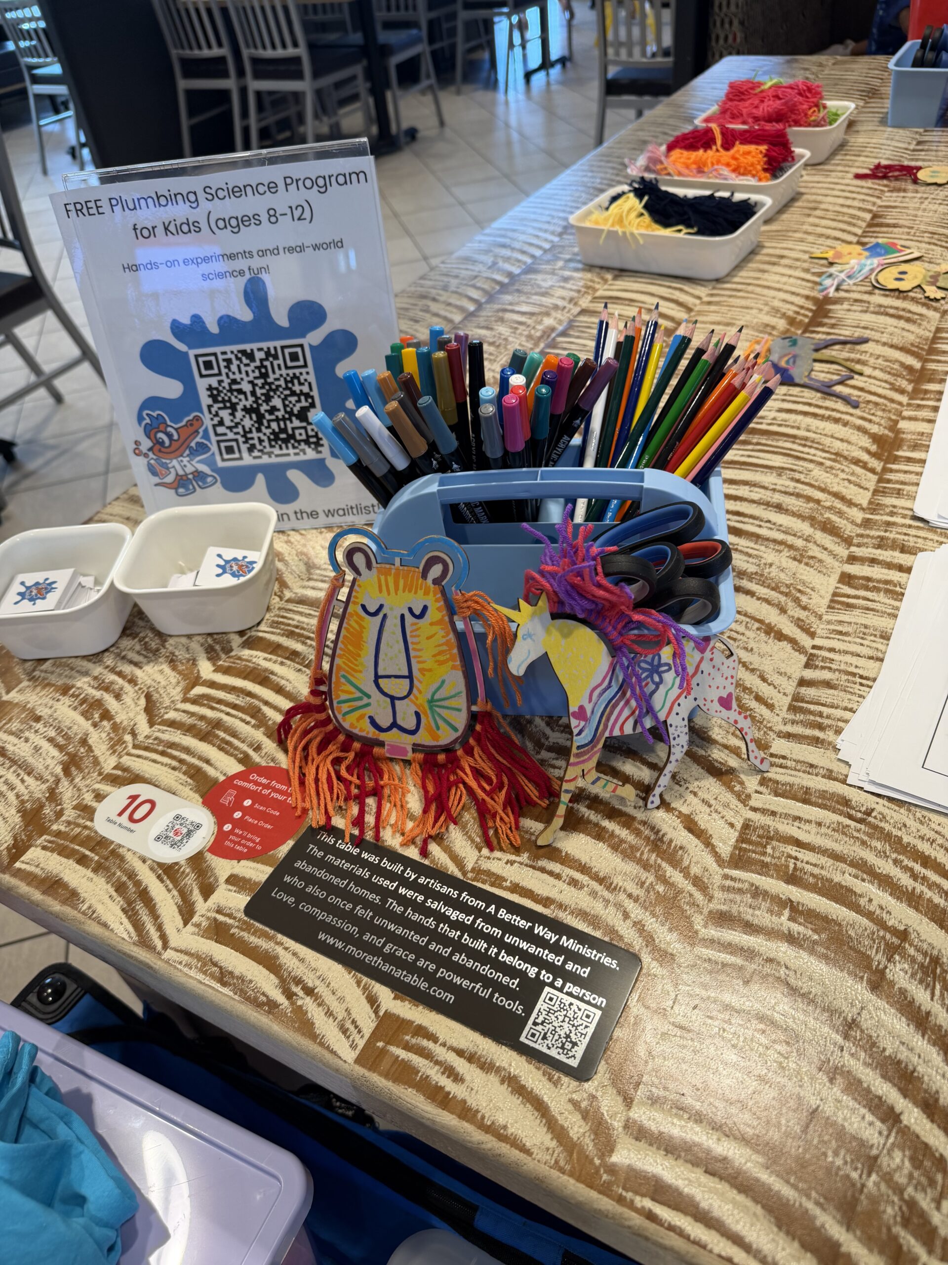 A sign reading Free Plumbing Science Program for Kids ages 8 to 12 beside markers, colored pencils, and craft projects on a table