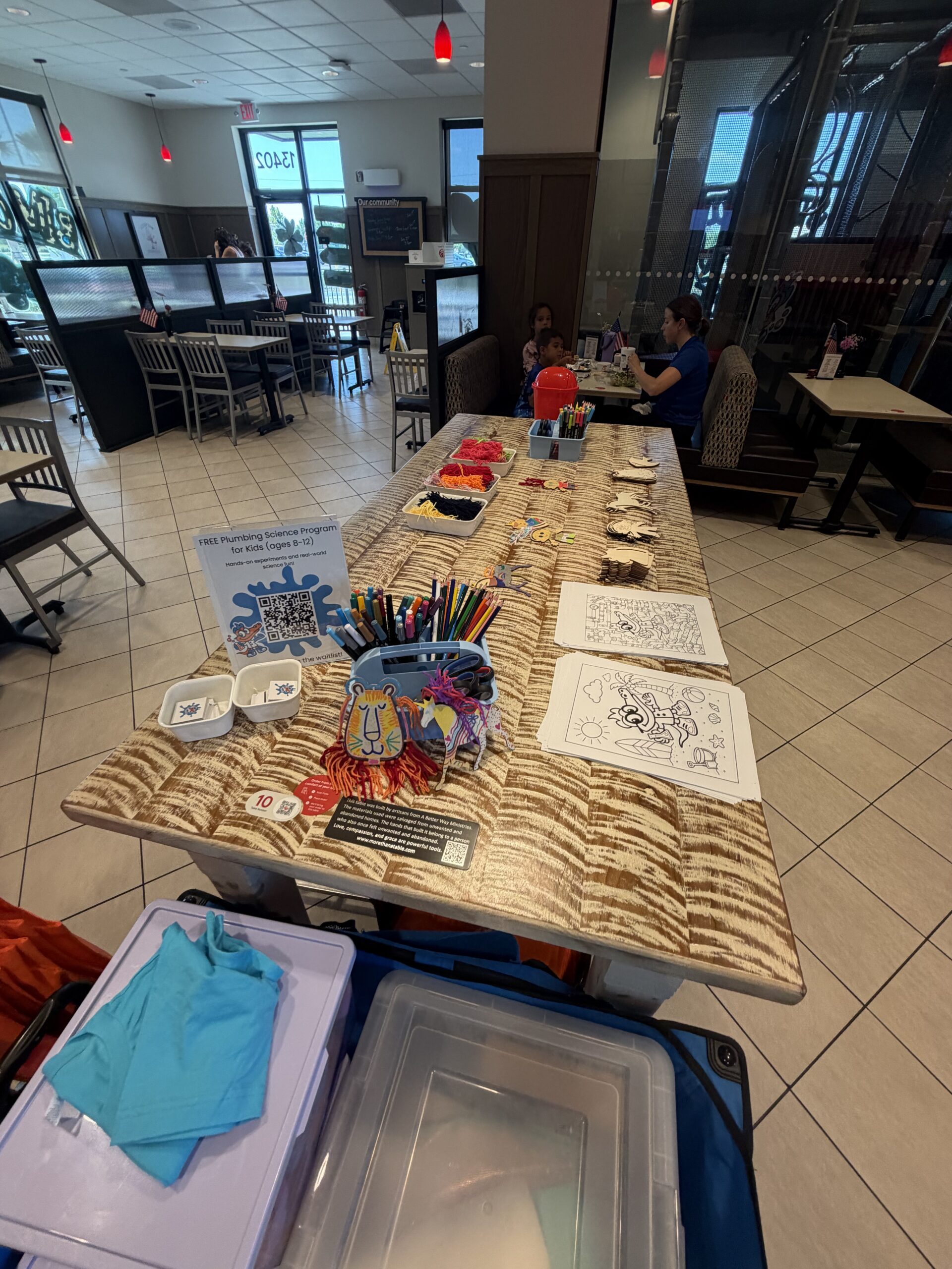A community event craft table with coloring pages, markers, and a Free Plumbing Science Program sign inside a restaurant