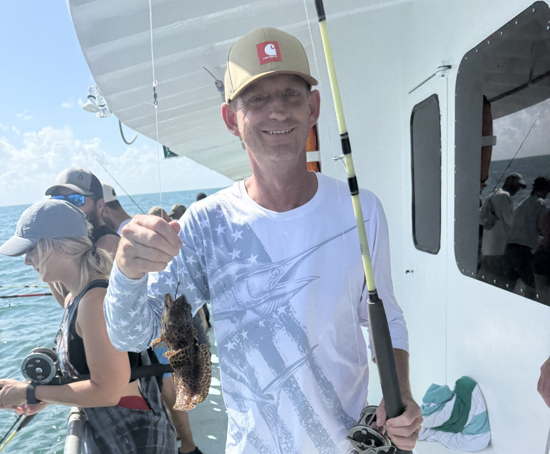 Smiling man on a boat holds up a small fish he just caught, with other anglers and the ocean in the background.