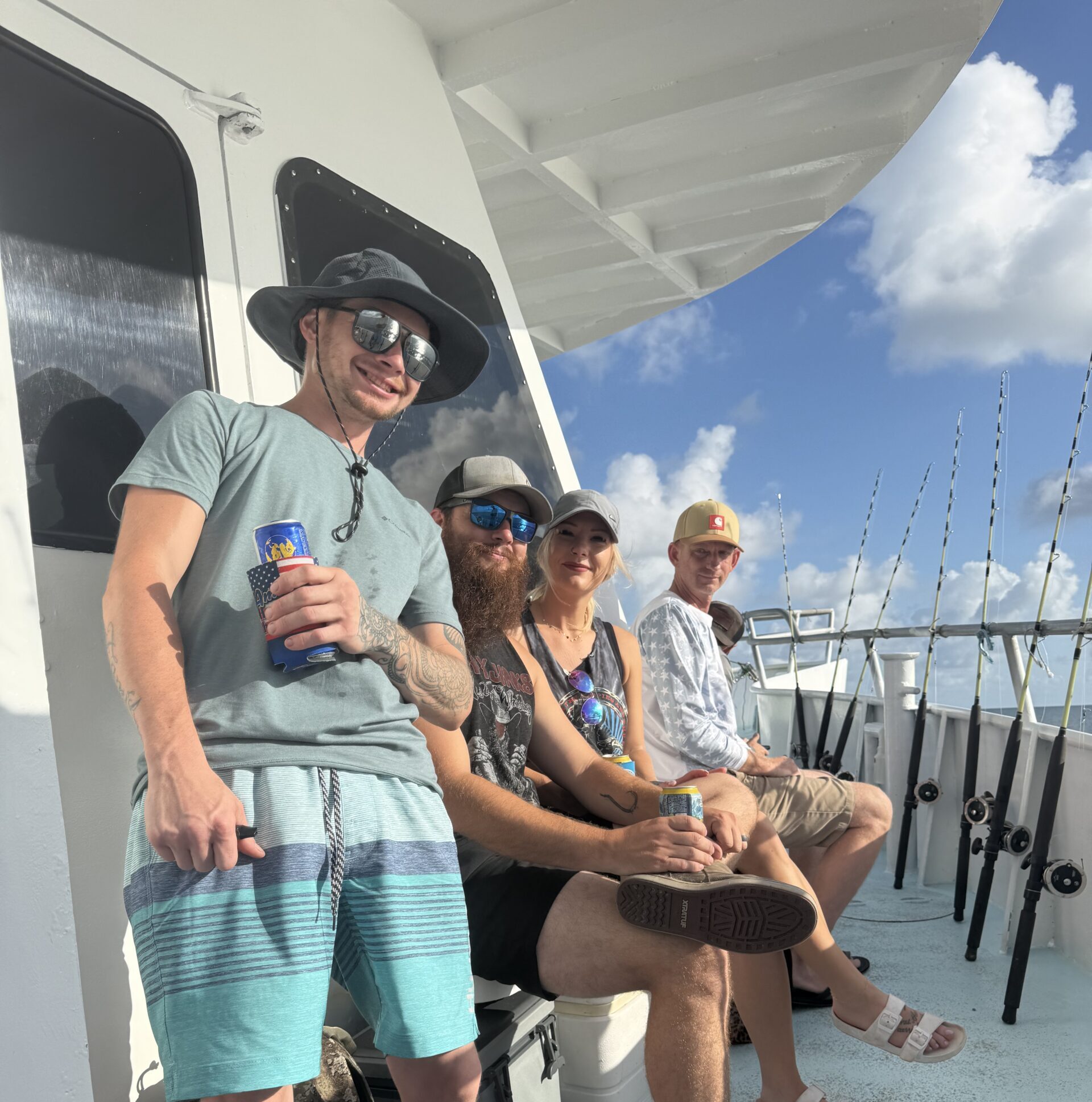 Four friends sit on a fishing boat deck, smiling at the camera, holding drinks with fishing rods lined up on the right side under a blue sky.