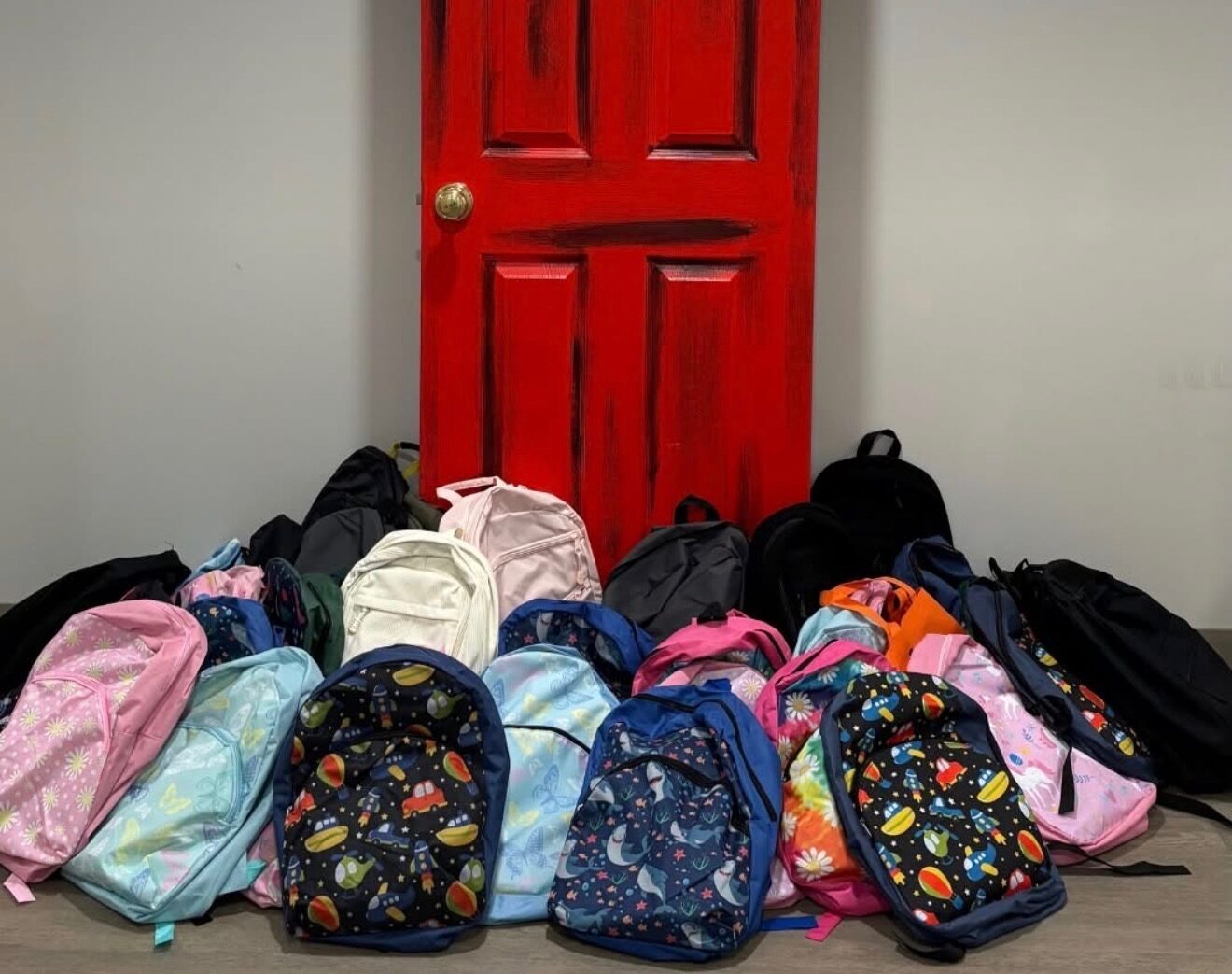 Thank You message and cartoon alligator mascot over a red door with a pile of colorful backpacks in front, likely a donation drive.